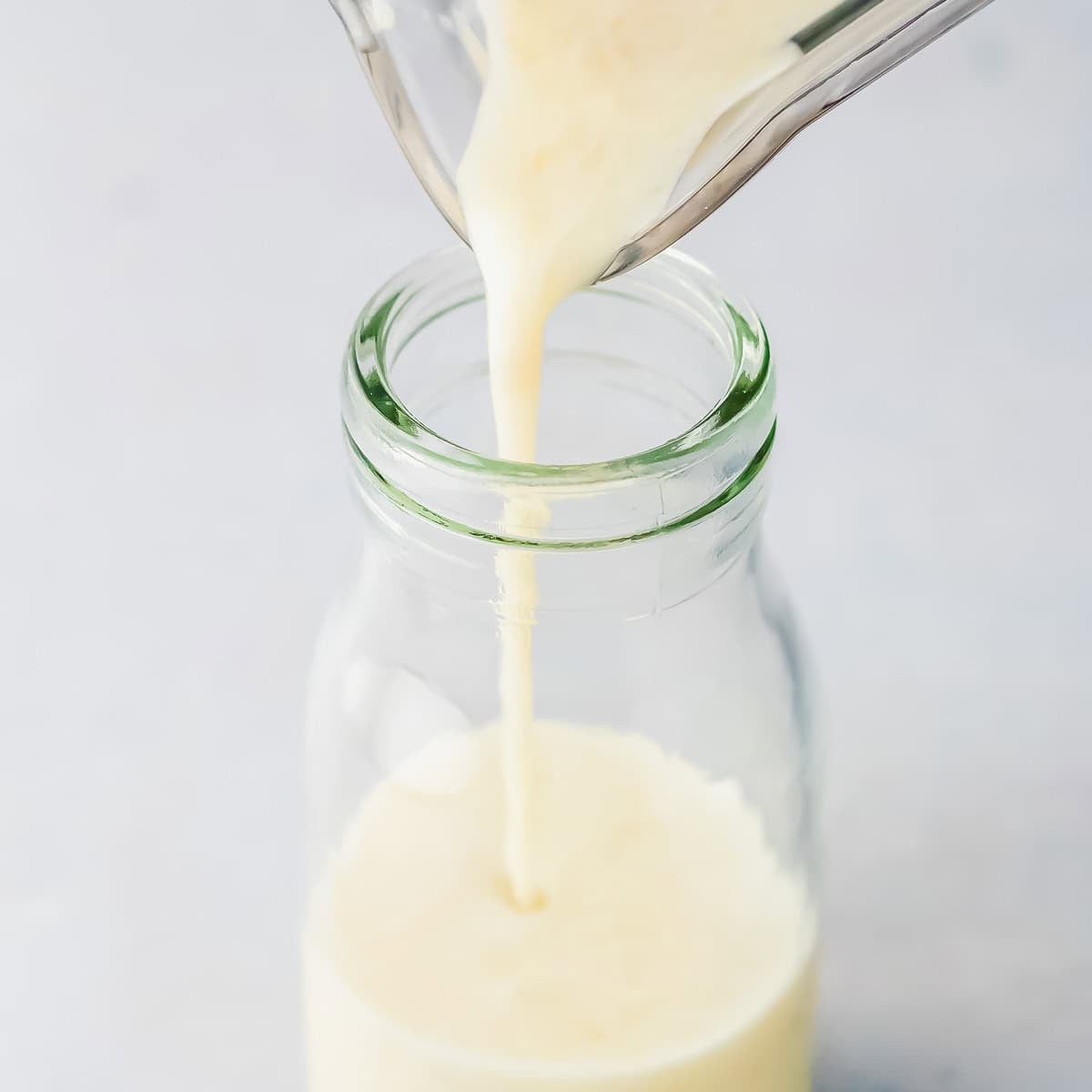 Creamy banana milk being poured from a blender into a clear glass bottle, filling it nearly to the top, against a light background.