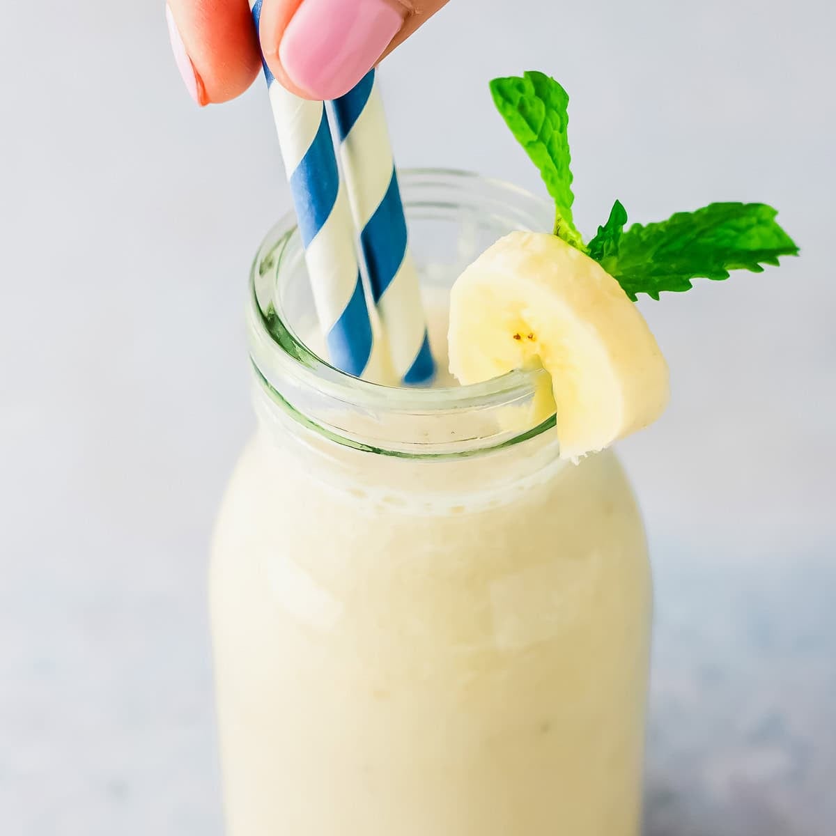 A hand with pink nail polish holds two blue-and-white striped straws in a glass jar of creamy banana milk, garnished with a banana slice and a sprig of mint.