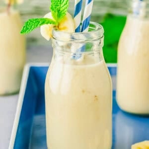 A glass bottle filled with creamy milk, garnished with a banana slice and mint sprig. Two striped straws are in the bottle, which is placed on a blue tray with another smoothie in the background.