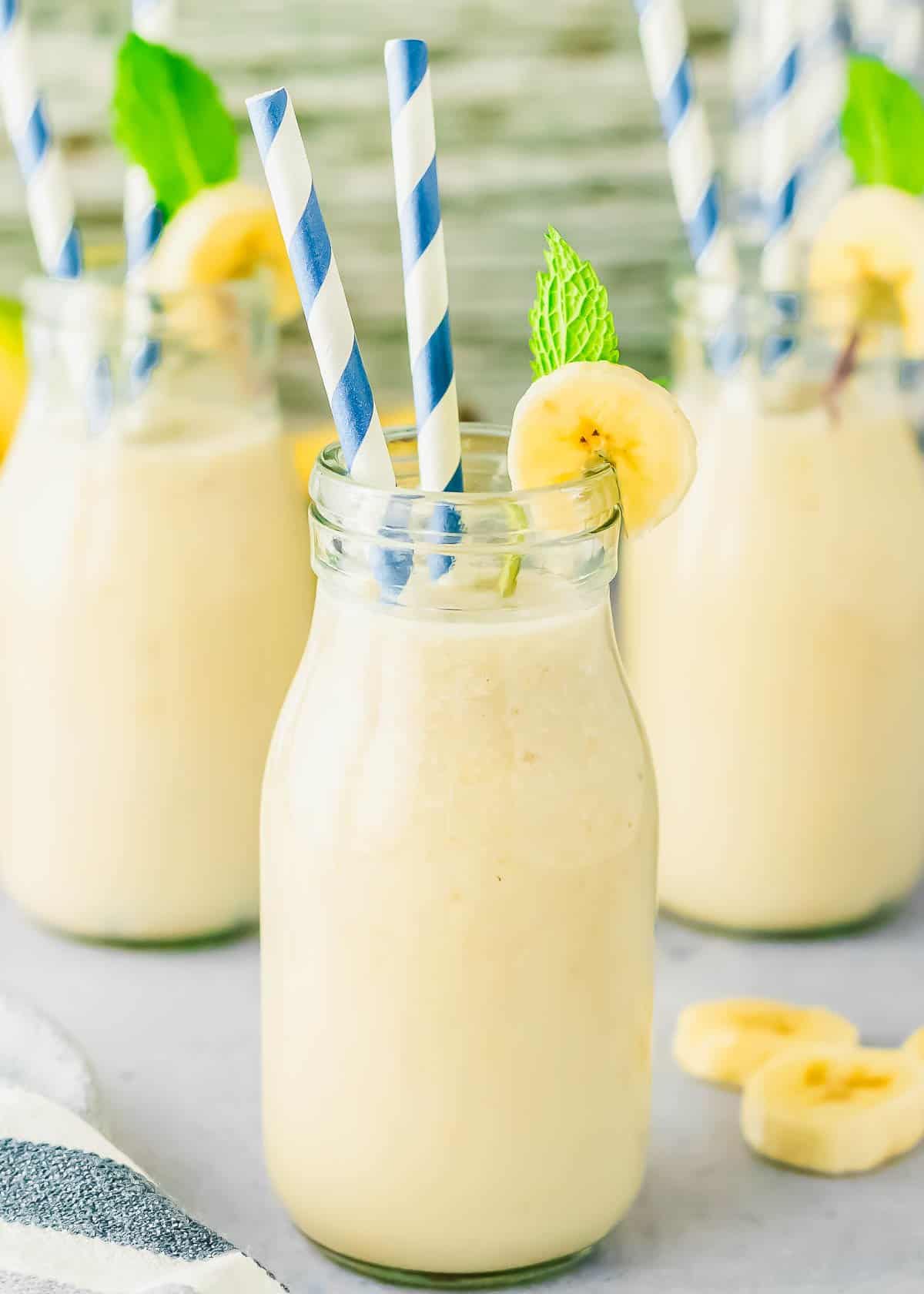 A glass bottle filled with Korean banana milk, garnished with a banana slice and mint, with a blue-striped straw. Three similar smoothie bottles appear in the background.