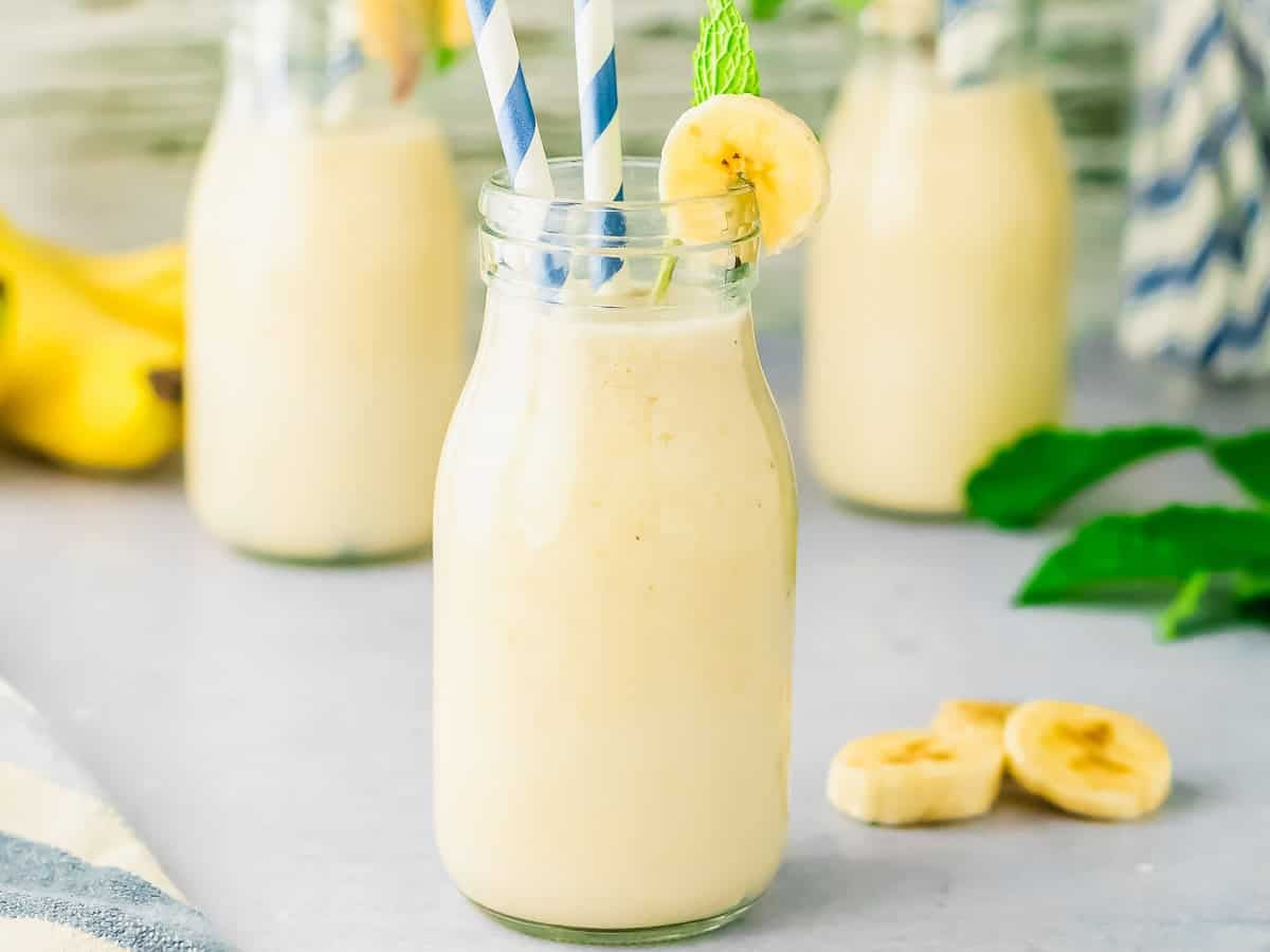 A glass bottle filled with banana milk, garnished with a banana slice and mint leaf, with two blue-striped straws. Two more bottles and banana slices are blurred in the background.