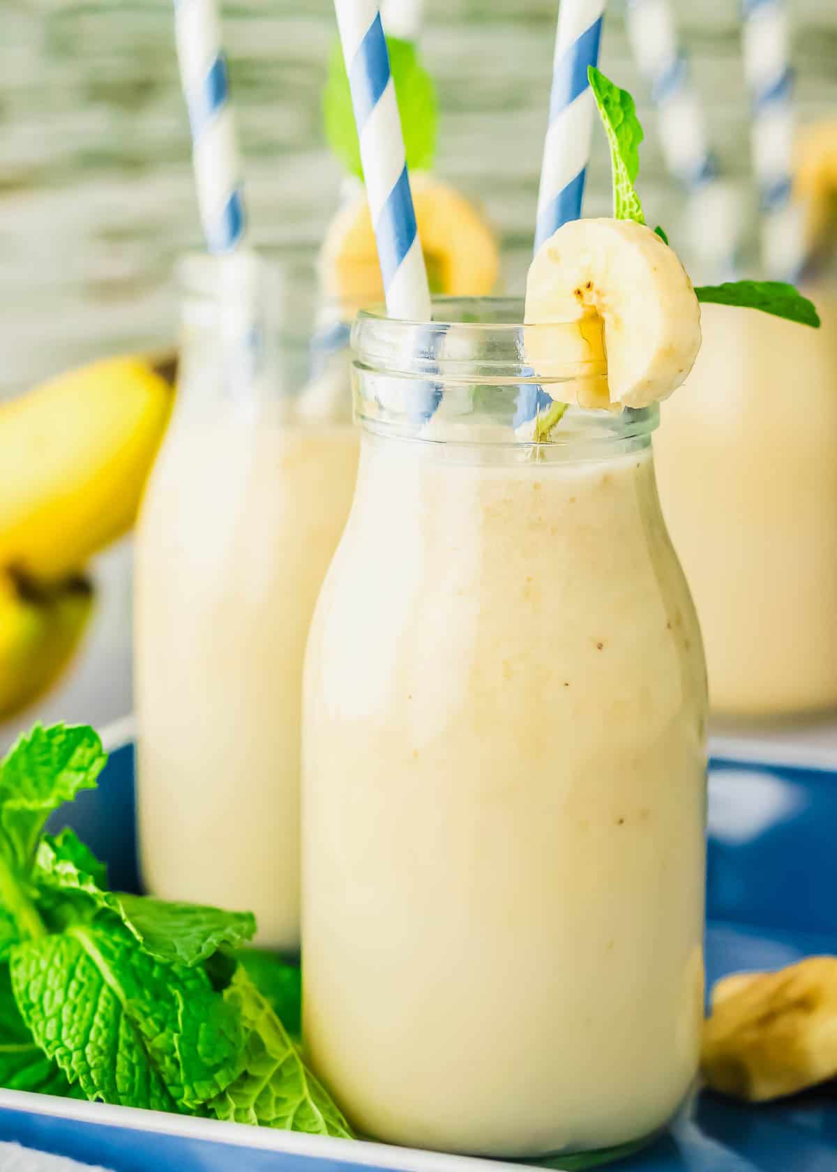A glass bottle of creamy banana drink with a blue striped straw, garnished with a banana slice and mint leaf. Two more drinks and fresh mint leaves are in the background.