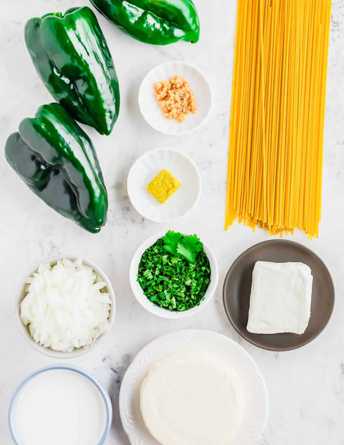 Ingredients on a white surface: three poblano peppers, uncooked spaghetti, chopped onion, chopped cilantro, minced garlic, a bouillon cube, cream cheese, a round cheese wheel, and a bowl of Mexican crema.