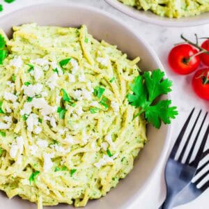 A bowl of creamy green pasta garnished with crumbled white cheese and fresh parsley, with a sprig of parsley and cluster of cherry tomatoes nearby. Black forks are placed beside the bowl on a white surface.