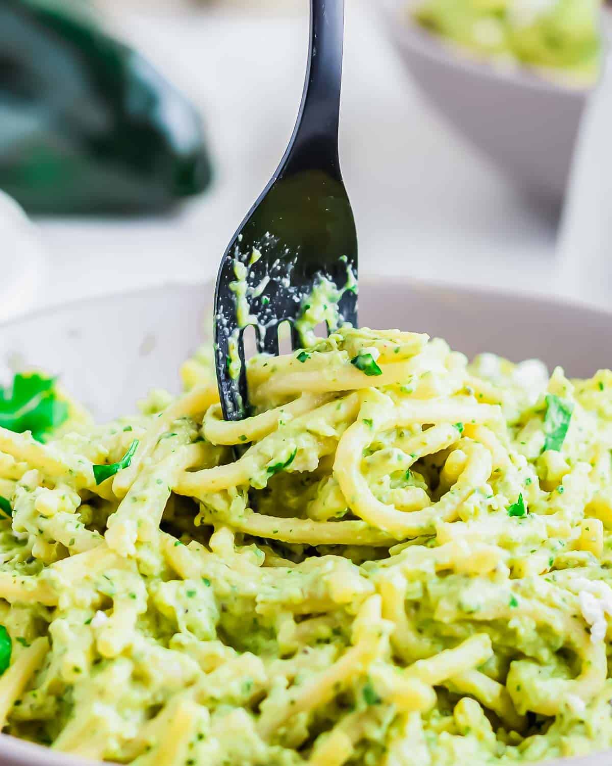 A close-up of creamy green pasta known as espagueti verde, being twirled by a black fork in a bowl. Fresh herbs are visible throughout the dish.