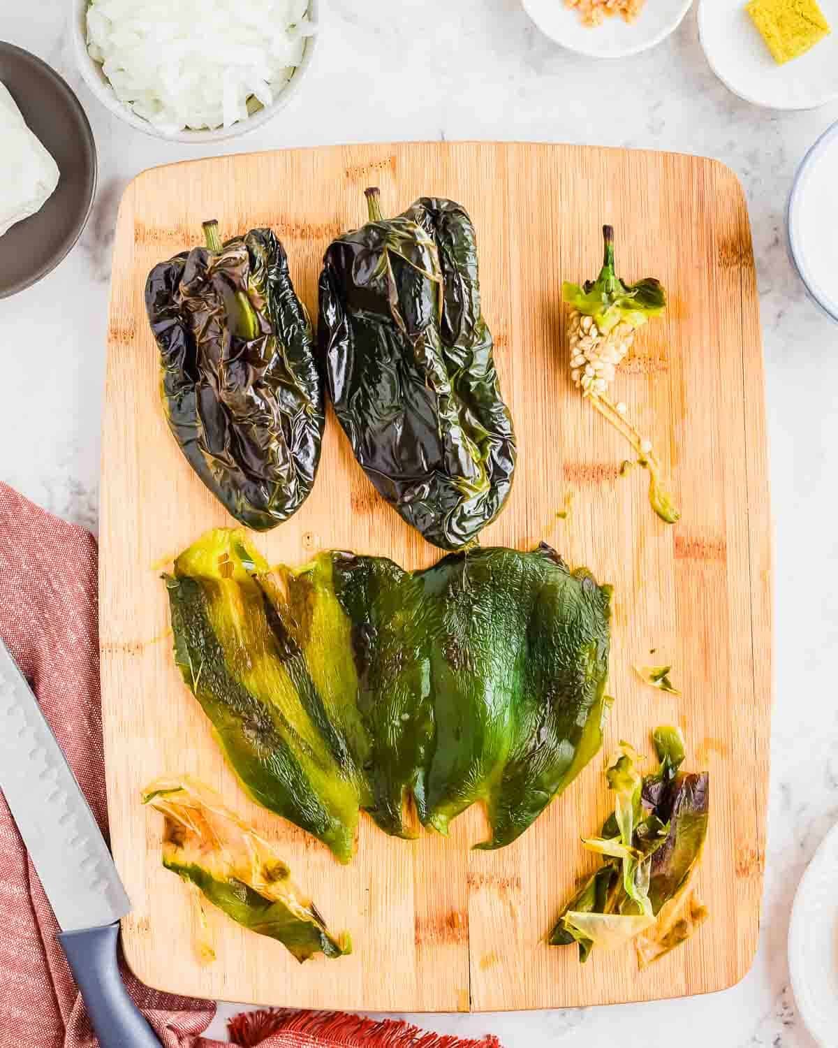 A wooden cutting board with roasted poblano peppers, some whole, some peeled and cut open, with skins and seeds removed. A knife and small bowls with ingredients are nearby on a white surface.