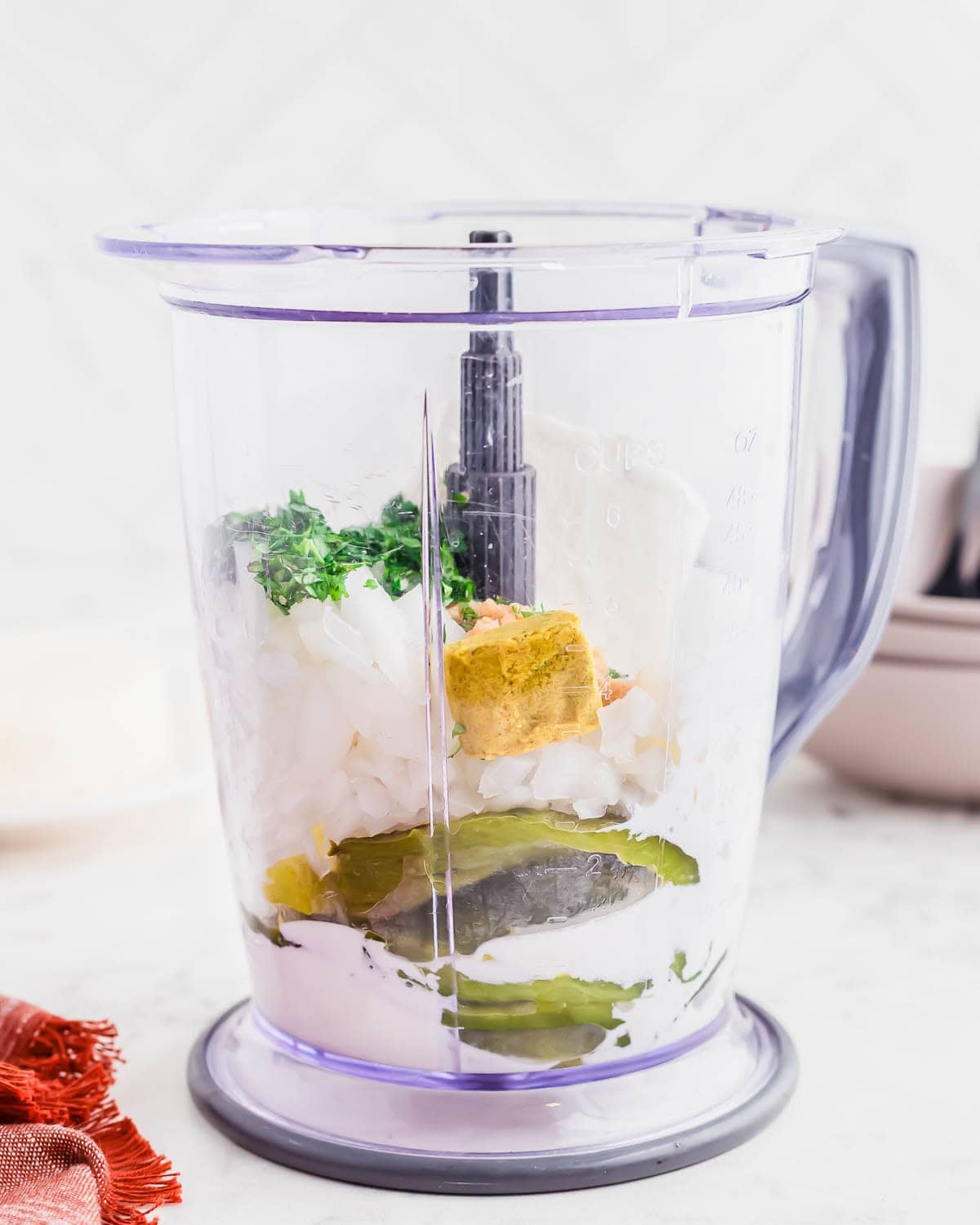 A clear blender cup filled with chopped onions, roasted poblano peppers, herbs, a yellow bouillon cube, and white creamy ingredients, sitting on a white counter.