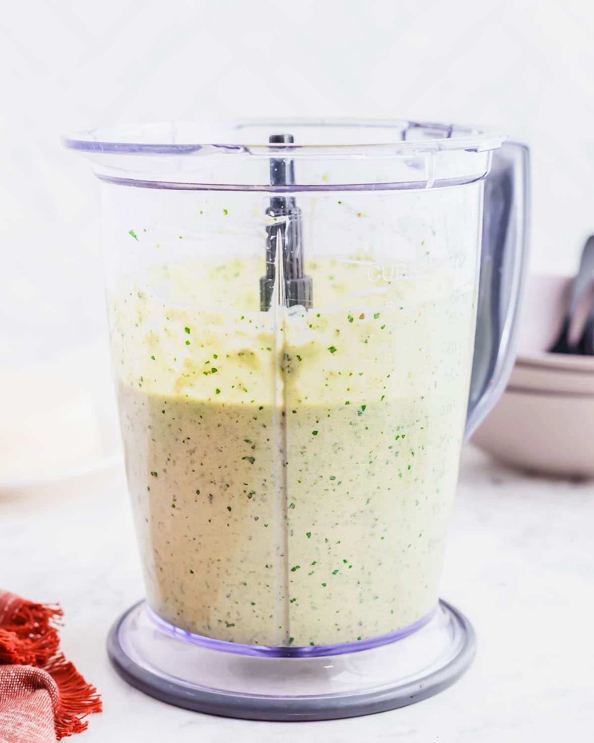 A clear blender pitcher filled with a creamy, verde sauce mixture speckled with herbs sits on a white countertop. Stacks of white bowls and a red cloth are in the background.