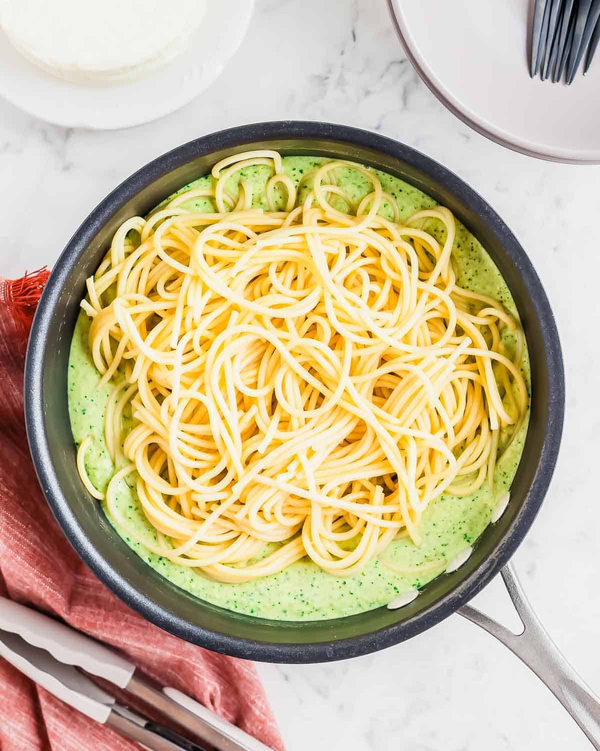 A pan filled with cooked spaghetti on top of a green sauce, placed on a marble surface with a red cloth, tongs, and white plates with black forks nearby.
