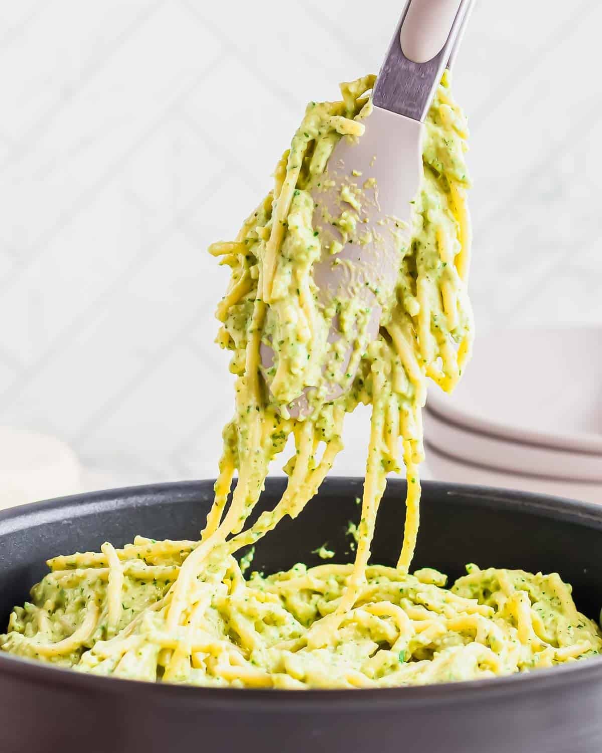 A utensil lifts a serving of spaghetti coated in a creamy green poblano sauce from a black pan, with several stacked plates blurred in the background.