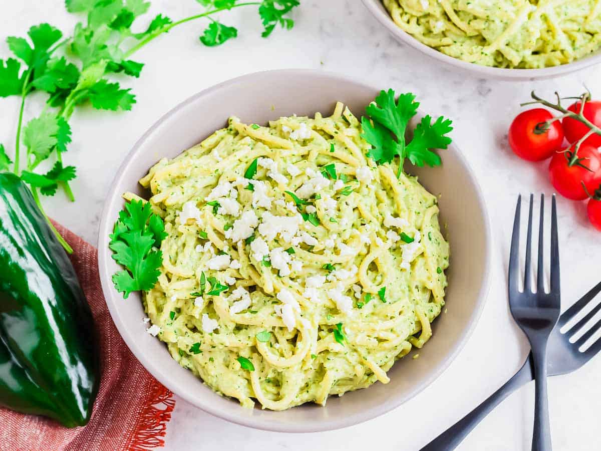 A bowl of creamy espagueti verde garnished with crumbled cheese and fresh cilantro, surrounded by another bowl of pasta, cherry tomatoes, cilantro sprigs, a poblano pepper, and two black forks on a white surface.