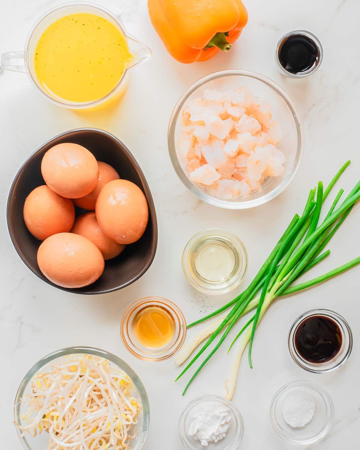A top-down view of cooking ingredients on a white surface, including eggs, shrimp, yellow bell pepper, green onions, bean sprouts, broth, and various sauces in small bowls.