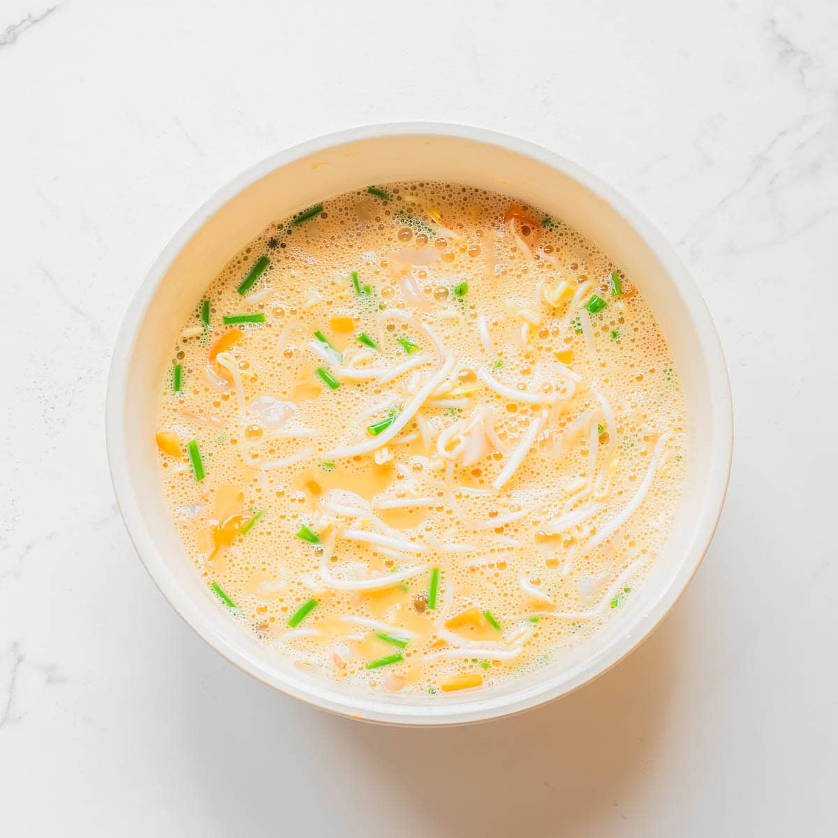 A white bowl filled with a beaten egg mixture containing chopped green onions and bean sprouts, placed on a white marble surface.