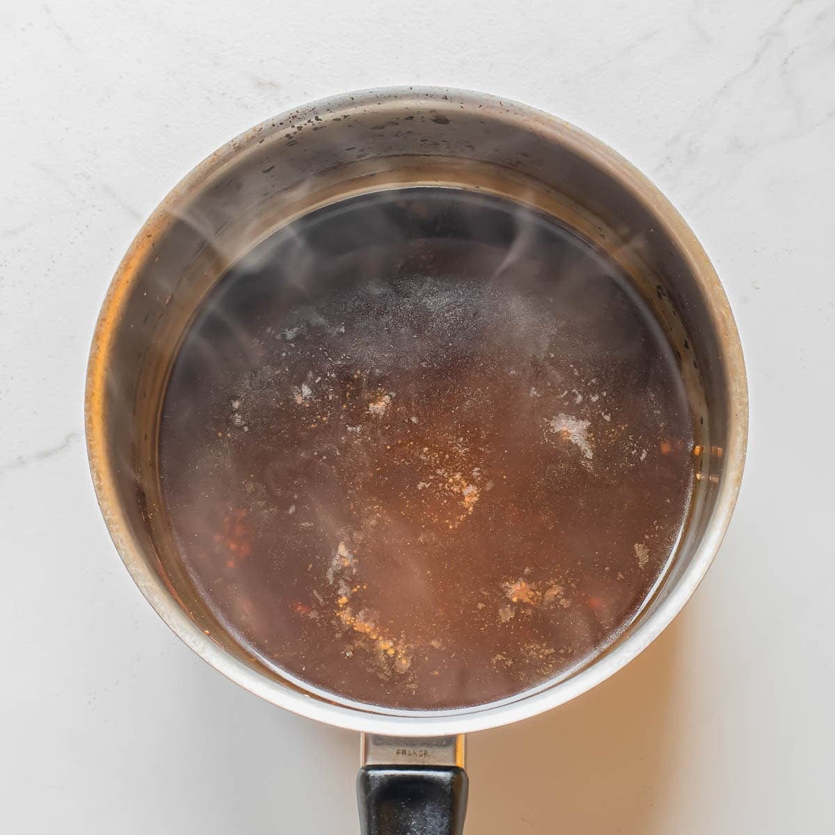 A stainless steel saucepan filled with steaming dark brown sauce, viewed from above, sitting on a white marble surface.