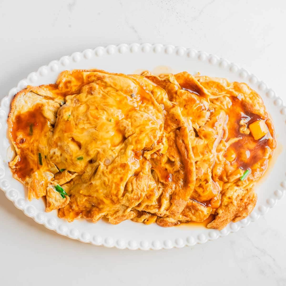 A white oval plate with several pieces of egg foo young with shrimp topped with a glossy, amber-colored sauce and garnished with small green onions on a light surface.