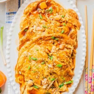 A platter of shrimp egg foo young topped with brown sauce, chopped green onions, sesame seeds, and small shrimp, served on a white oval dish with chopsticks and garnishes on the side.