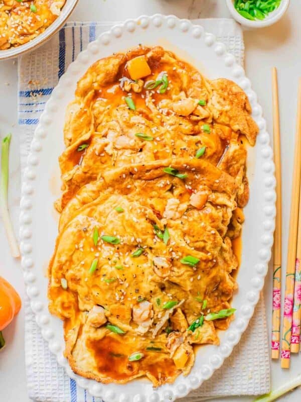 A platter of shrimp egg foo young topped with brown sauce, chopped green onions, sesame seeds, and small shrimp, served on a white oval dish with chopsticks and garnishes on the side.