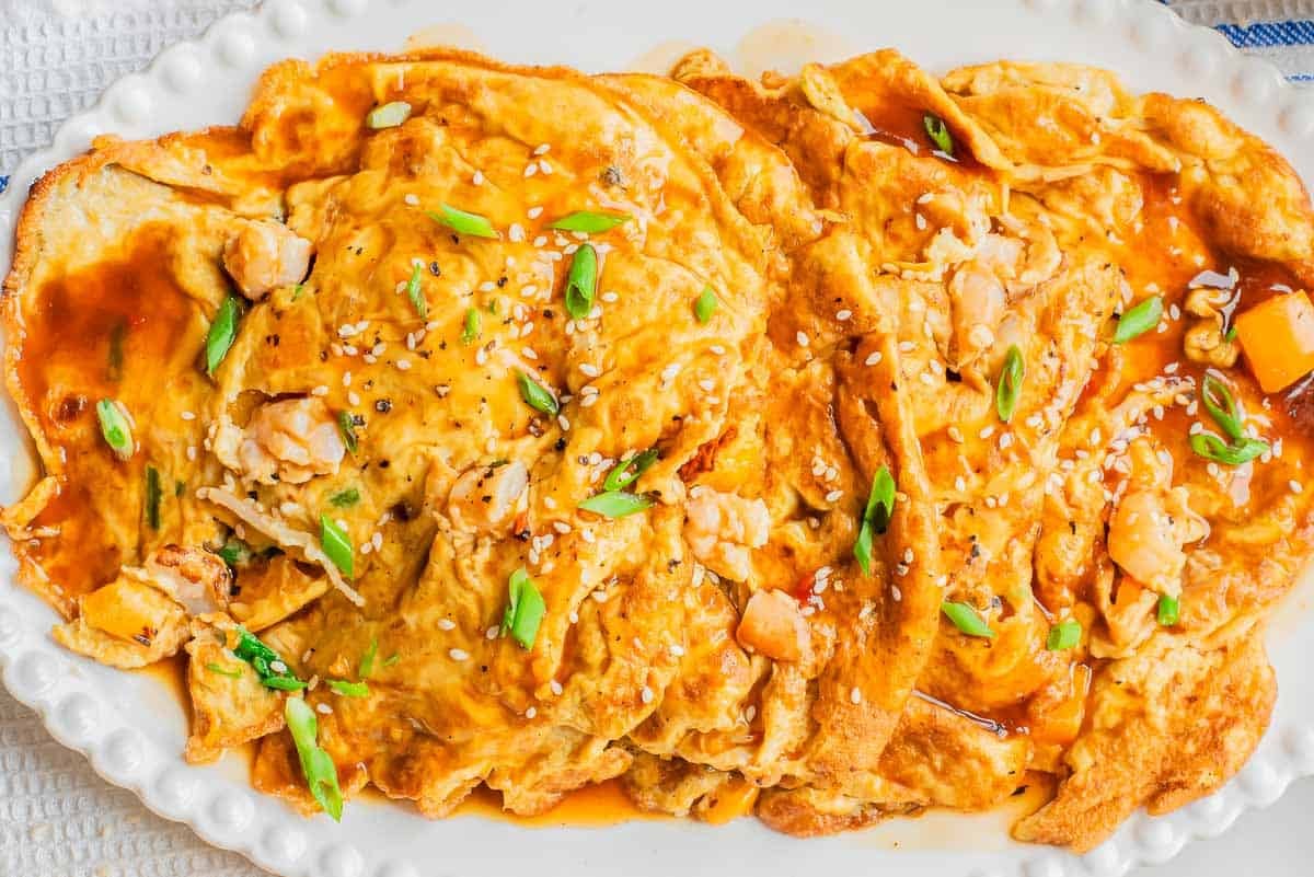 A close-up of a platter filled with golden brown Chinese omelettes topped with a savory sauce, chopped green onions, sesame seeds, and bits of chicken. The omelette is arranged on a white, scalloped-edged plate.