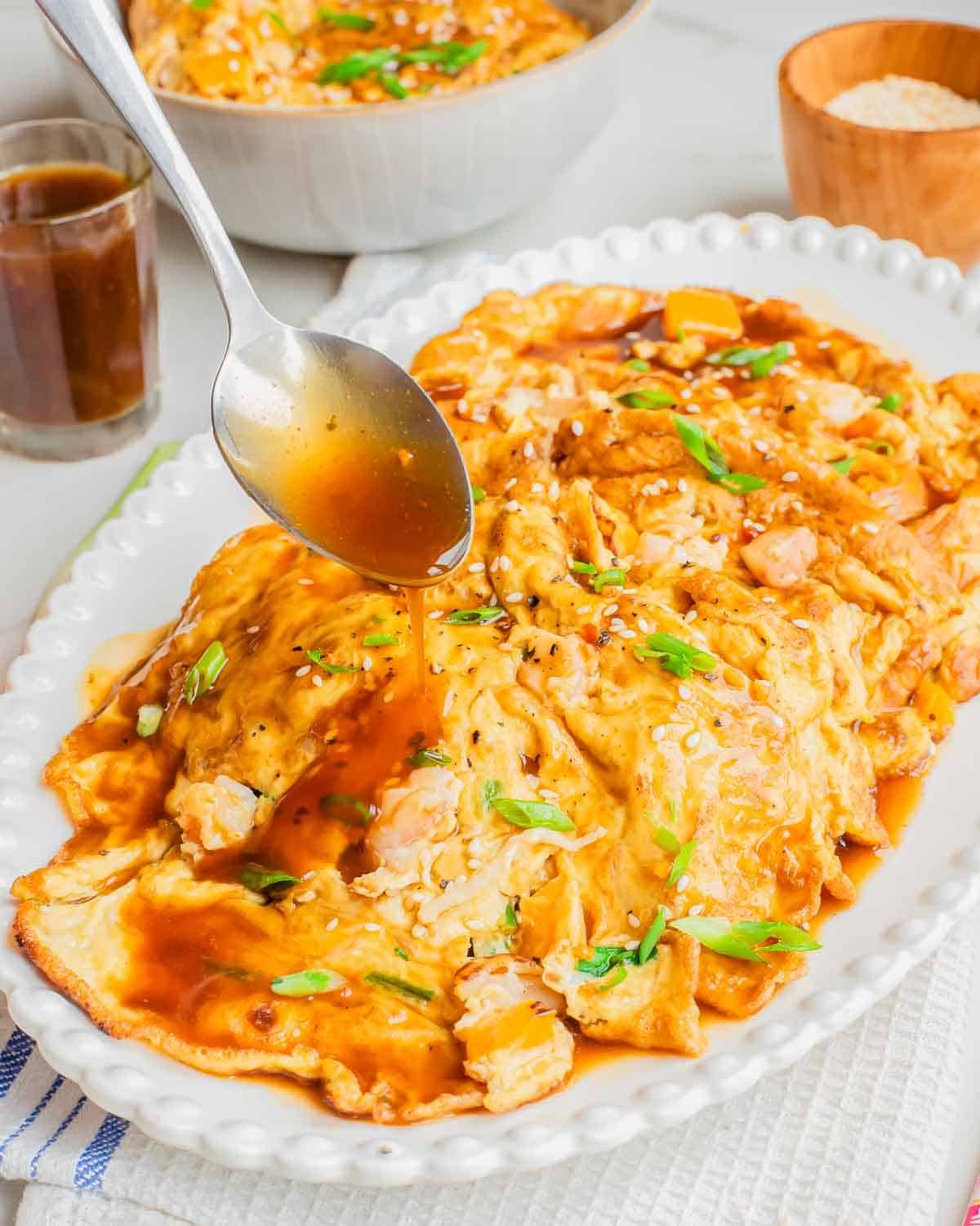 A spoon pours brown sauce over egg foo young shrimp garnished with green onions and sesame seeds, served on a white oval dish.