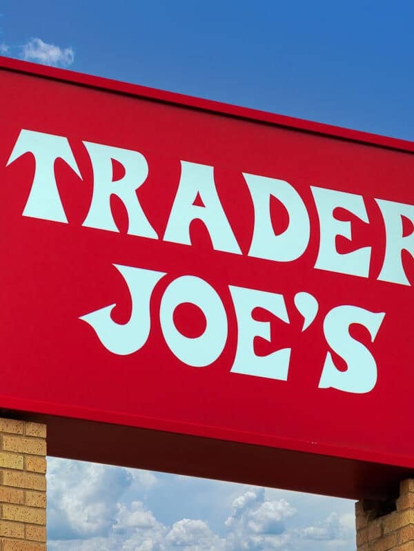A large red Trader Joe’s sign with bold white letters is mounted on a brick base, set against a blue sky with scattered clouds.