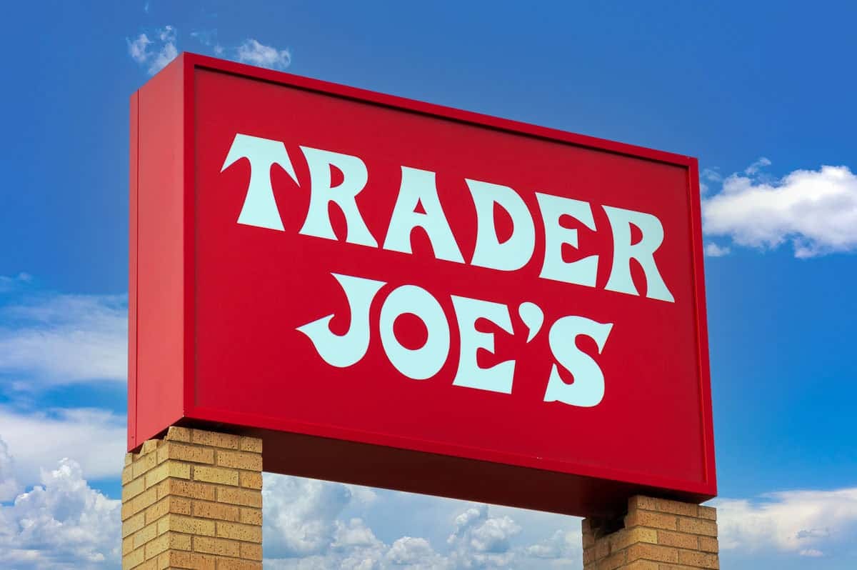 A large red Trader Joe&rsquo;s sign with bold white letters is mounted on a brick base, set against a blue sky with scattered clouds.