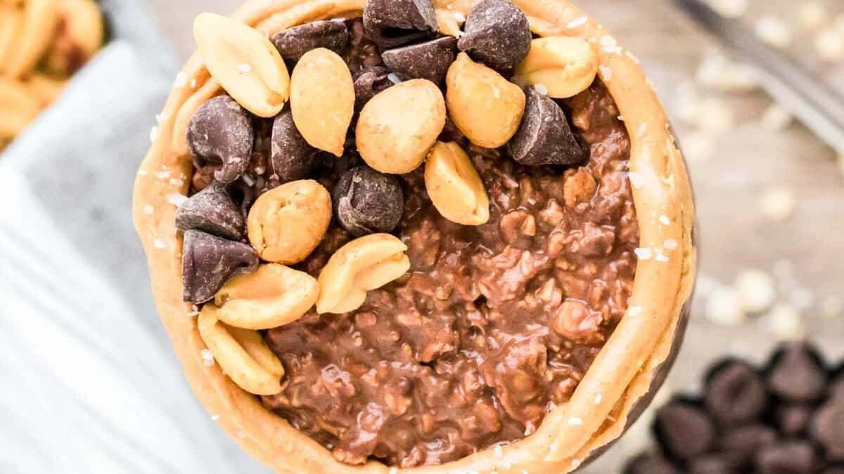 A close-up of a bowl filled with chocolate oatmeal, topped with chocolate chips and roasted peanuts, and surrounded by a layer of creamy peanut butter on the rim.