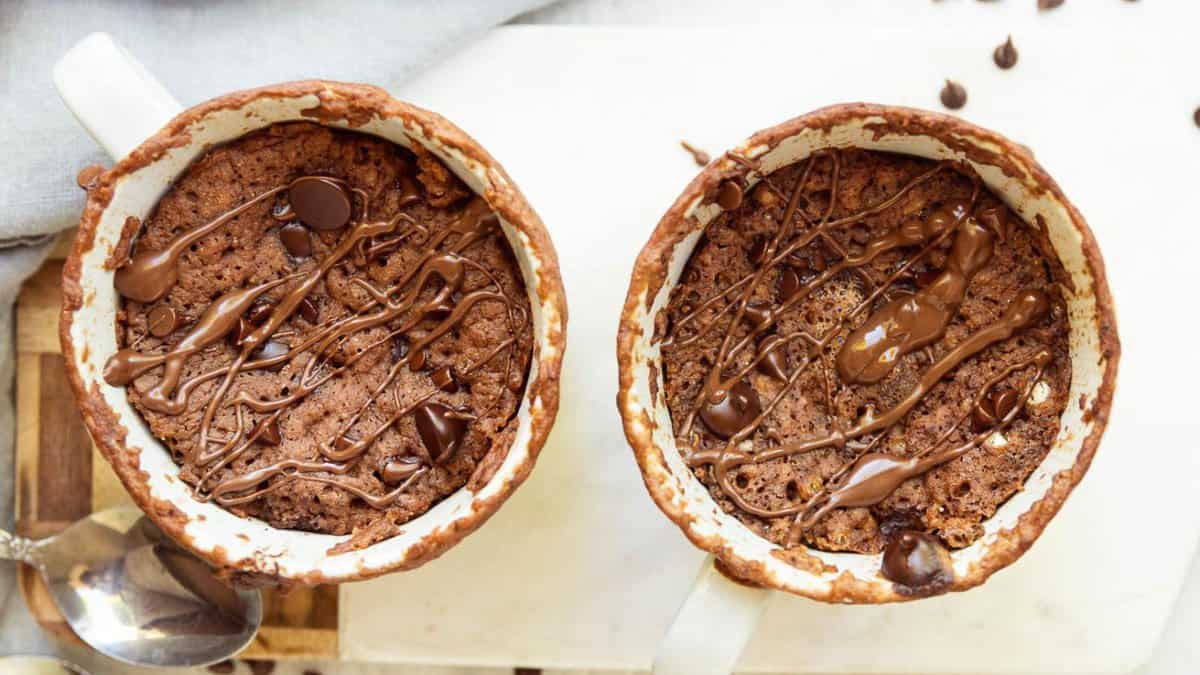 Two chocolate mug cakes in white mugs, each topped with melted chocolate drizzle and chocolate chips, sit side by side on a light-colored surface with a spoon nearby.