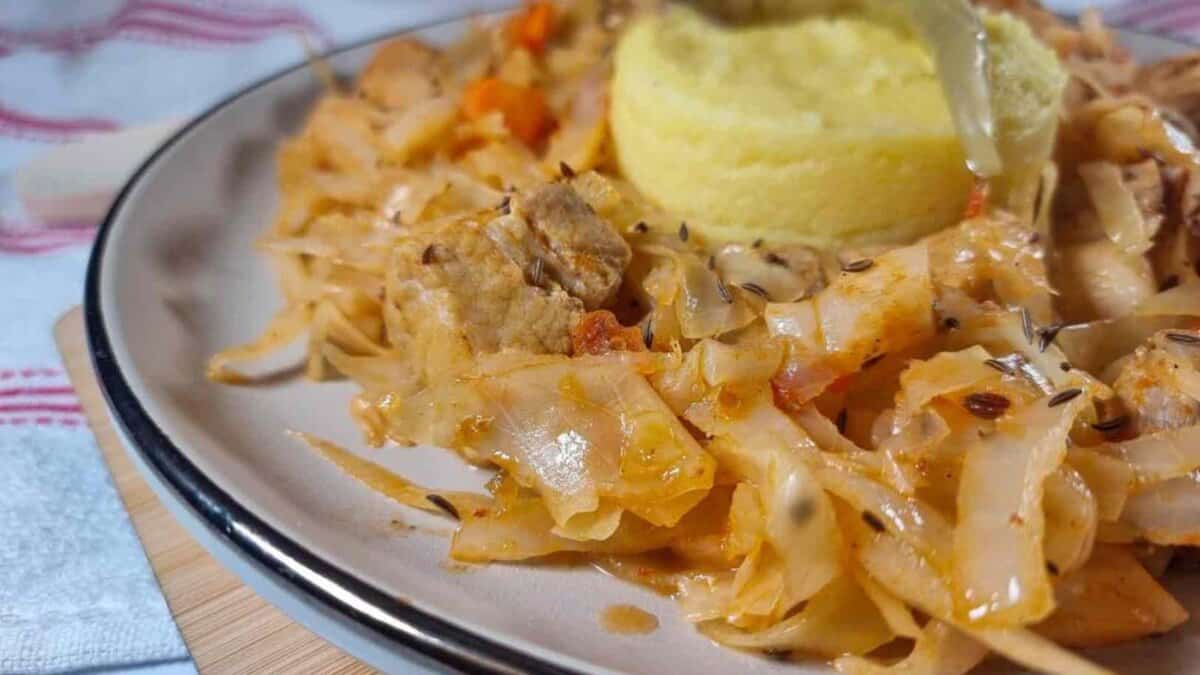 A close-up of a plate with stewed cabbage and pieces of meat, garnished with caraway seeds, served alongside a round portion of creamy yellow polenta.