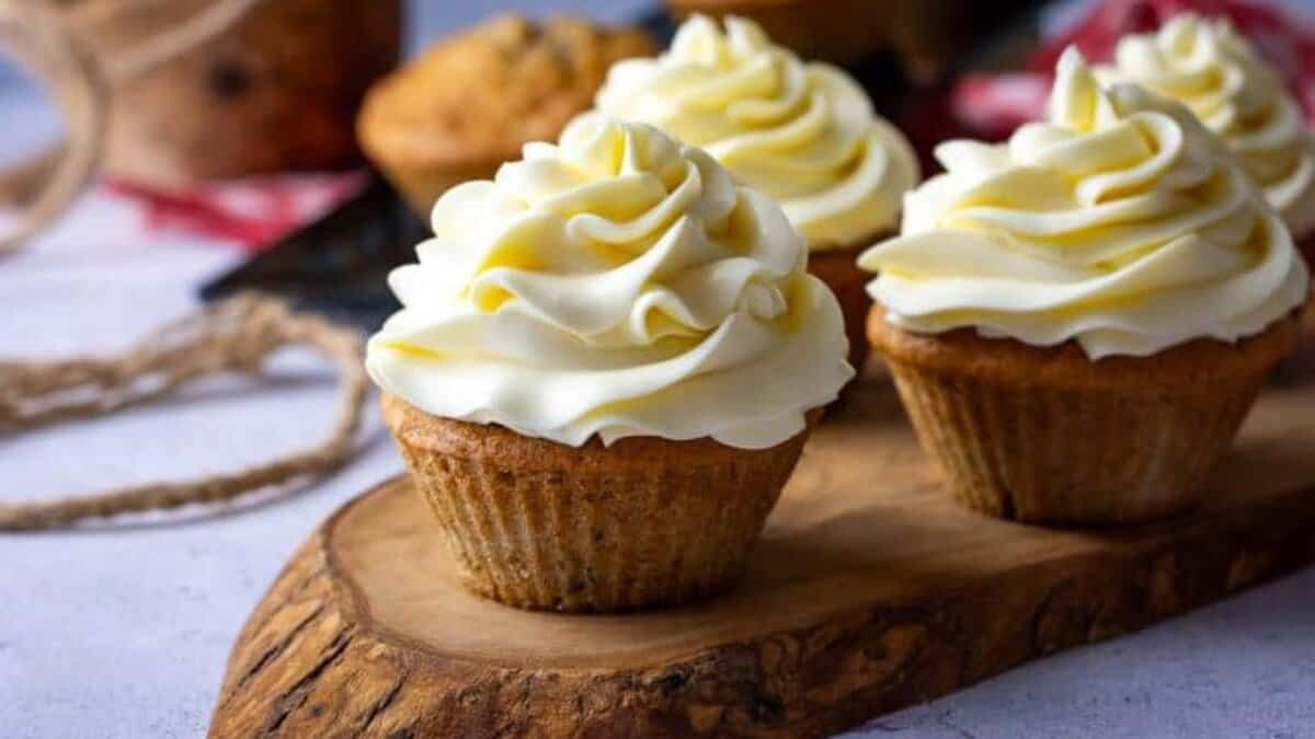 Three cupcakes with swirled vanilla frosting on top are displayed on a rustic wooden board, with a blurred background including another cupcake and some twine.