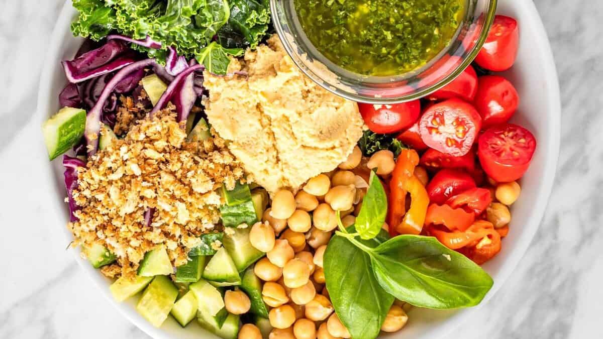 A bowl filled with fresh ingredients including kale, red cabbage, cucumber, cherry tomatoes, chickpeas, yellow peppers, hummus, bread crumbs, and a small bowl of green dressing, garnished with fresh basil leaves.
