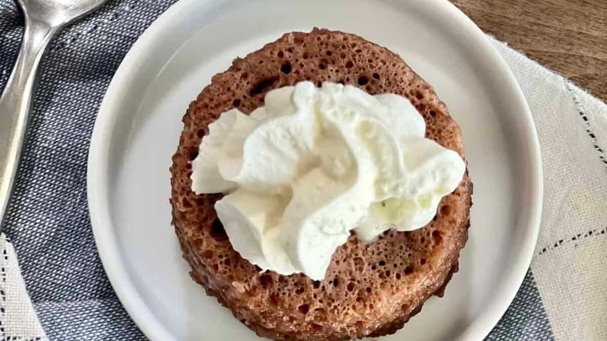 A chocolate mug cake topped with whipped cream is served on a white plate, next to a spoon and a gray-and-white napkin.