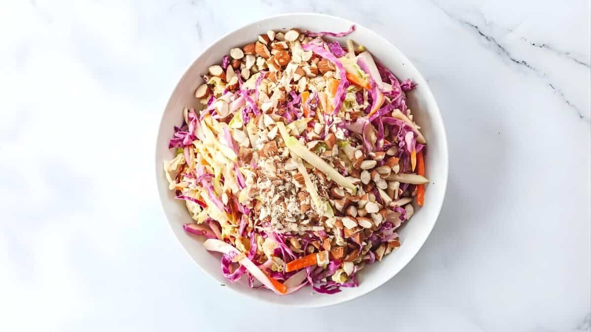 A white bowl filled with a colorful salad made of shredded cabbage, carrots, sliced almonds, and sunflower seeds, placed on a white marble surface.