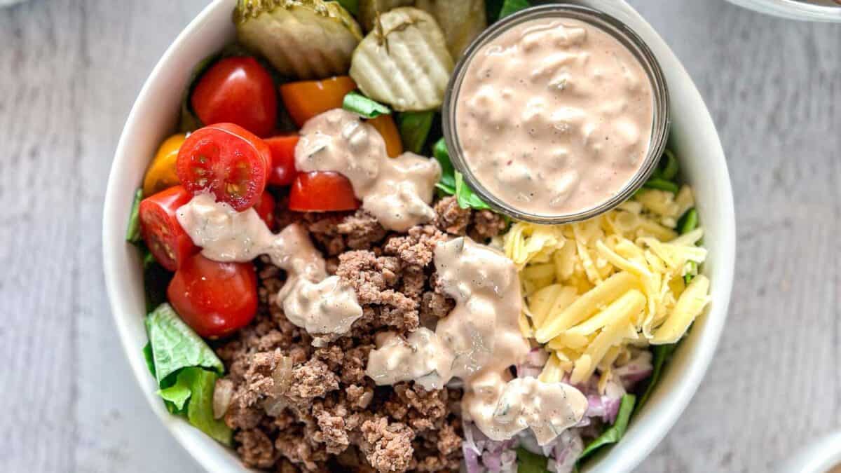 A bowl filled with ground beef, shredded cheese, sliced cherry tomatoes, pickles, chopped lettuce, and diced onions, topped with a creamy dressing, with an extra cup of dressing on the side.