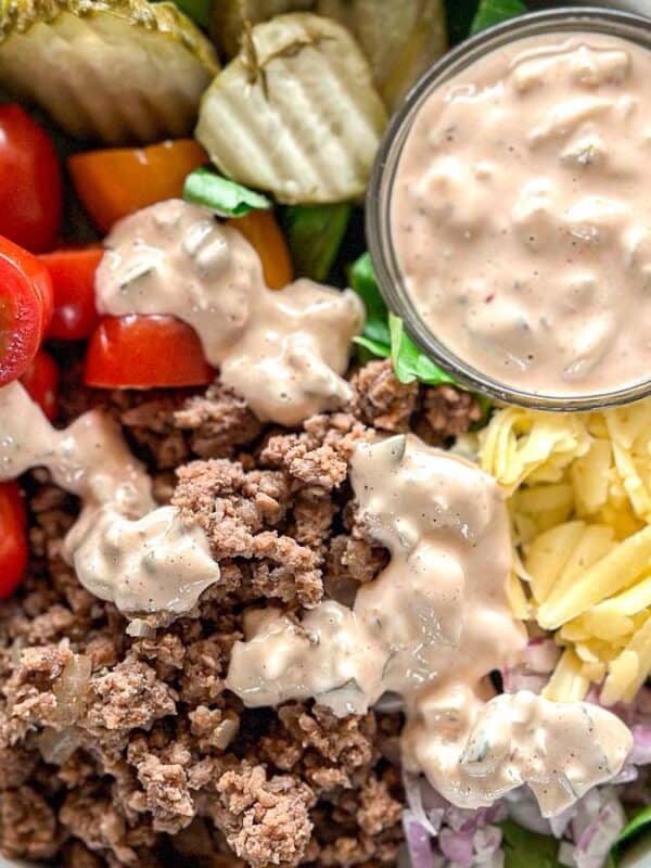 A bowl filled with ground beef, shredded cheese, sliced cherry tomatoes, pickles, chopped lettuce, and diced onions, topped with a creamy dressing, with an extra cup of dressing on the side.