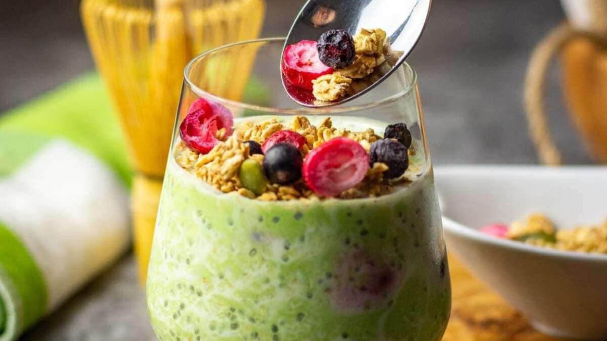 A glass filled with green chia pudding, topped with granola, sliced red berries, and blueberries. A spoon above the glass holds some granola and berries, ready to be added.