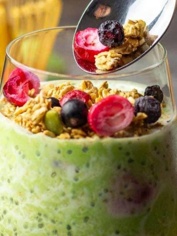 A glass filled with green chia pudding, topped with granola, sliced red berries, and blueberries. A spoon above the glass holds some granola and berries, ready to be added.