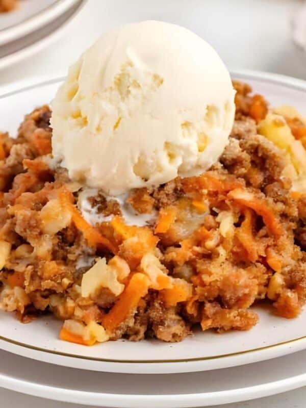 A serving of carrot cake crumble topped with a scoop of vanilla ice cream sits on a white plate, with forks, another plate, and a small bowl of chopped nuts in the background.