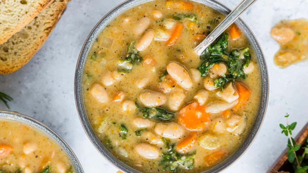 A bowl of creamy white bean soup with carrots and leafy greens, served with a spoon. Slices of bread are visible on the side. The soup appears hearty and thick.