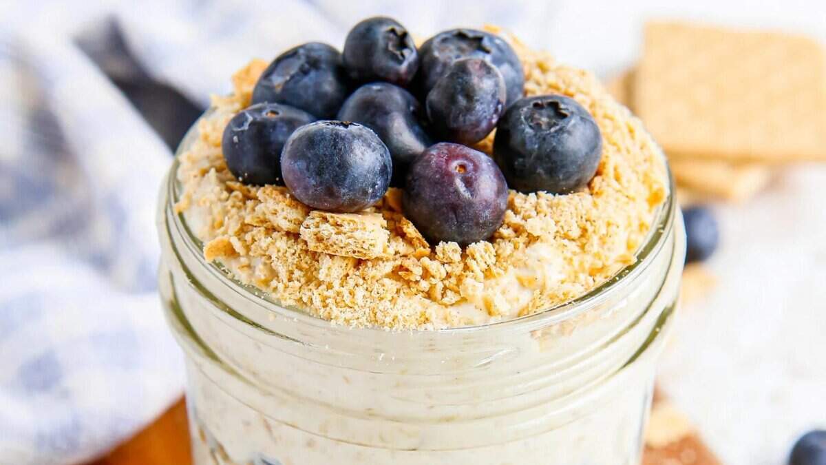 A mason jar filled with creamy oatmeal topped with crushed graham crackers and fresh blueberries, with a blurred background.