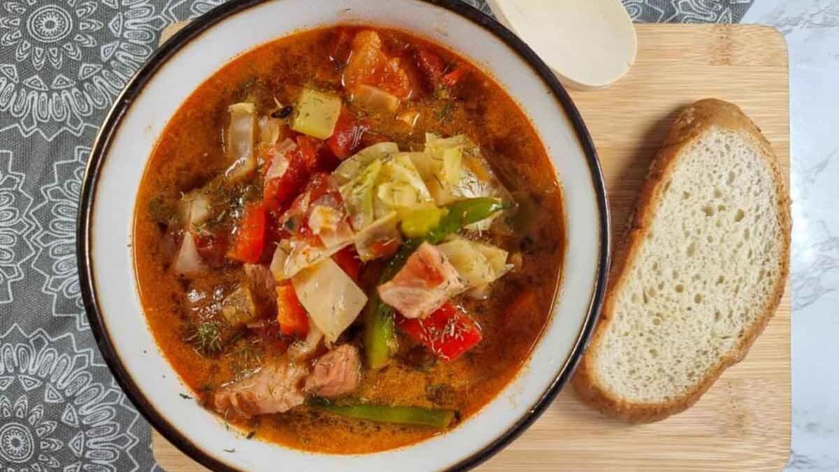 A bowl of vegetable and meat soup with chunks of cabbage, tomatoes, and green beans next to a slice of bread on a wooden board, with a white patterned tablecloth partially visible.
