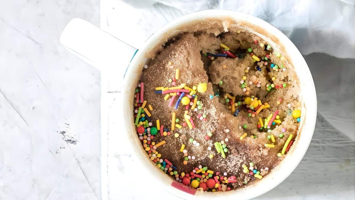 A mug cake topped with colorful sprinkles sits in a white mug on a light surface, with a portion scooped out from the side.