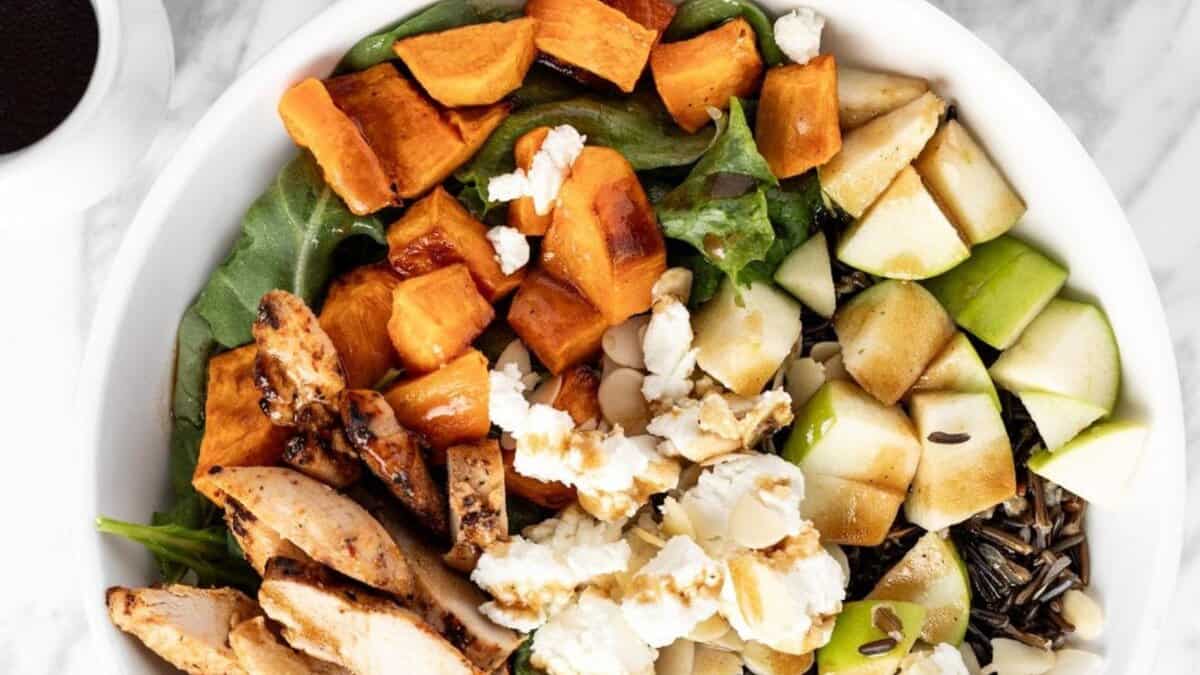 A salad bowl containing grilled chicken slices, roasted sweet potatoes, diced green apple, wild rice, crumbled cheese, sliced almonds, and mixed greens on a marble surface.