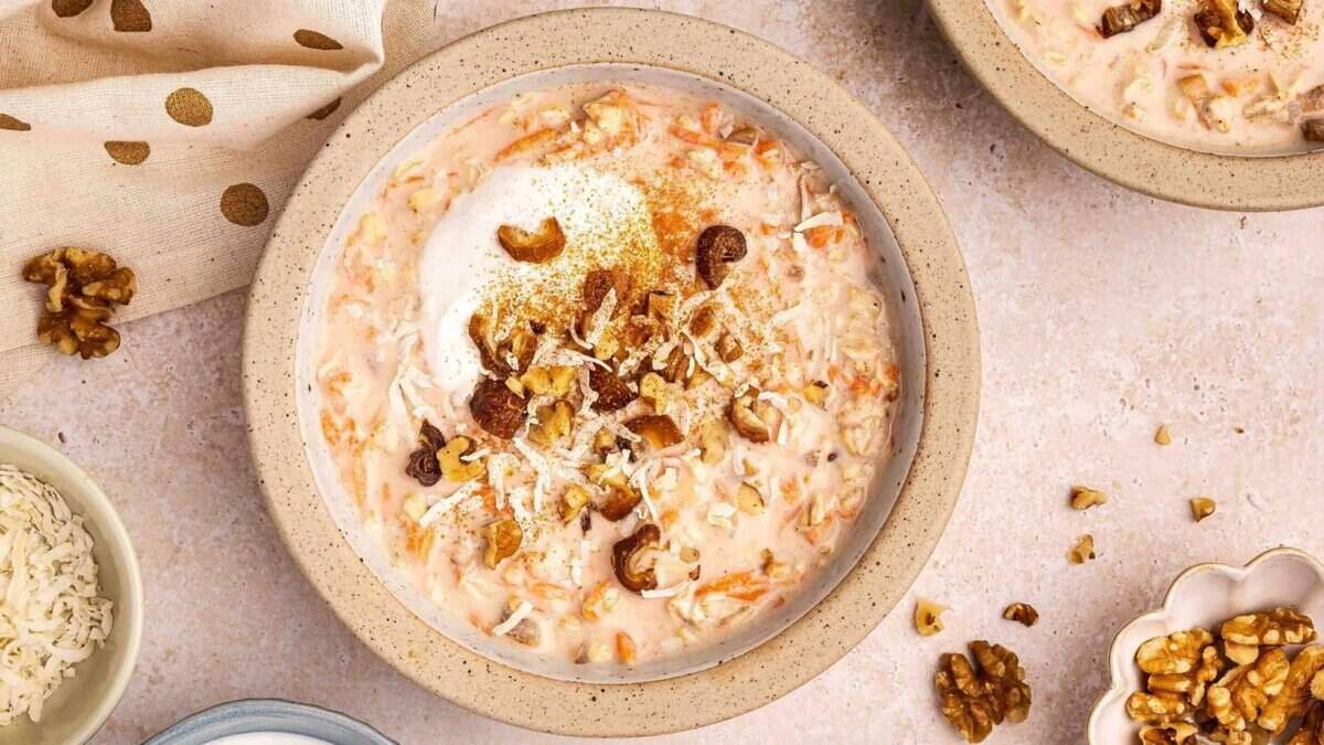 A bowl of creamy oatmeal topped with shredded coconut, chopped walnuts, and a sprinkle of spices, surrounded by small bowls of coconut and walnuts on a light-colored surface.