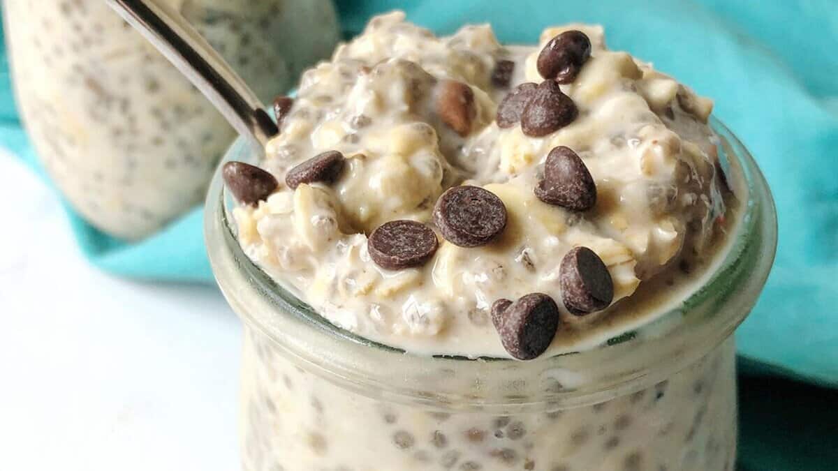 A close-up of a glass jar filled with creamy overnight oats topped with chocolate chips, with a spoon inside. A blue cloth is in the background.