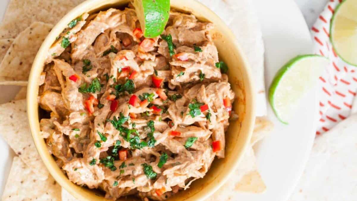 A bowl of creamy shredded chicken dip garnished with chopped cilantro and red peppers, served with tortilla chips on the side and a wedge of lime on top.