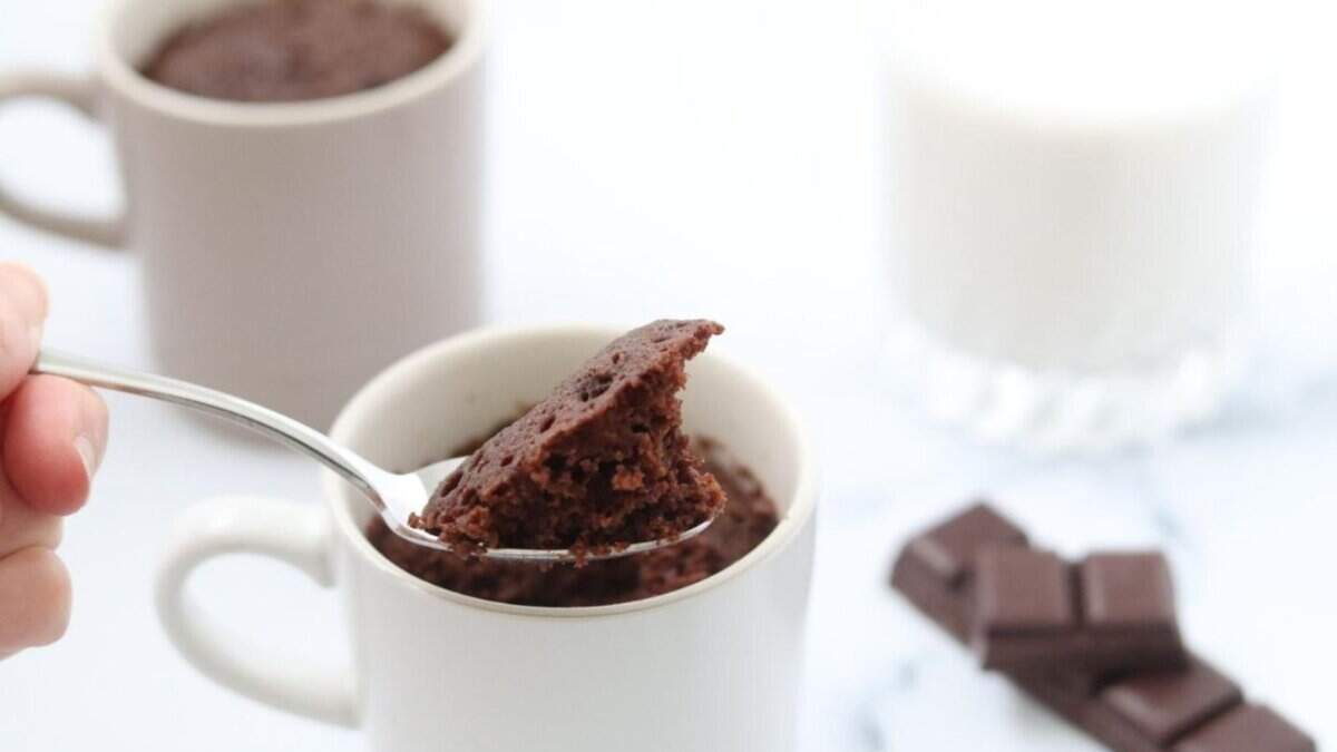 A close-up of a spoon holding a bite of chocolate mug cake above a white mug, with another mug, pieces of chocolate, and a glass of milk in the background.