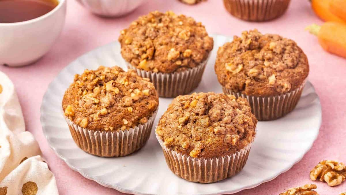Four carrot muffins with crumbly walnut topping sit on a white plate. A cup of tea, a muffin, and fresh carrots are partially visible in the background on a pink surface.