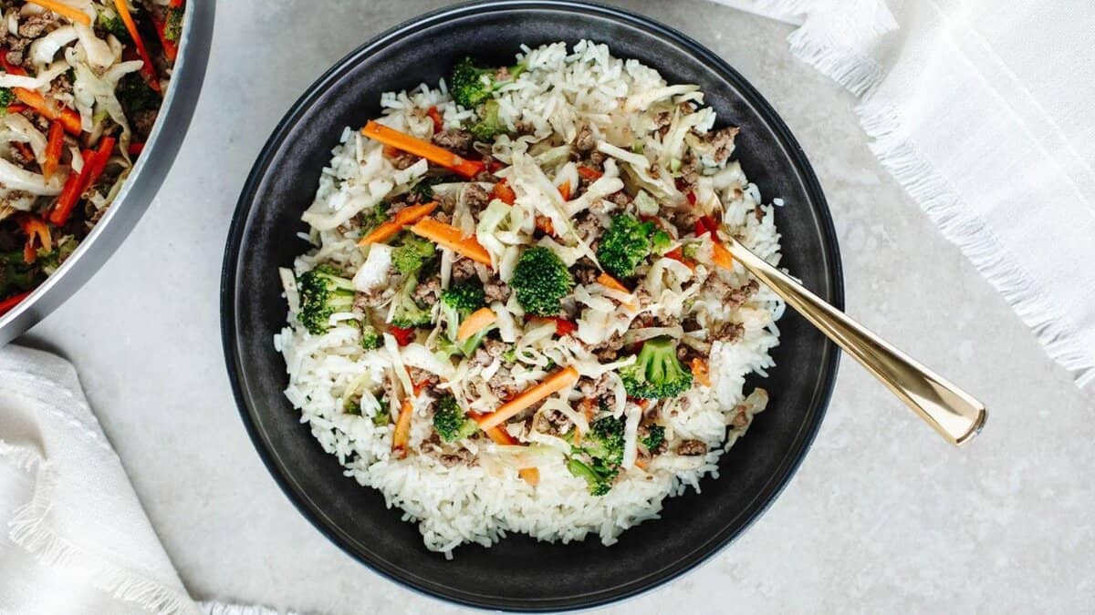 A black bowl filled with white rice, topped with ground meat, shredded cabbage, carrots, red bell pepper, and broccoli, sits on a light surface with a gold fork resting inside.