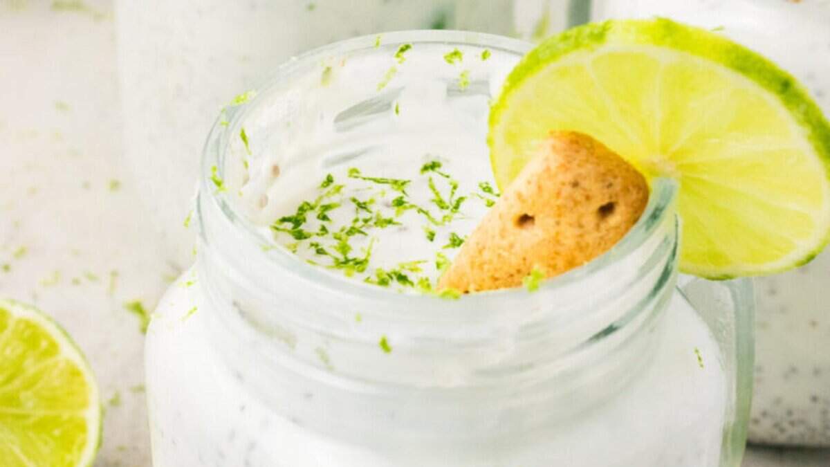 A mason jar filled with creamy white dessert, garnished with lime zest, a lime wheel, and a cookie on the rim. Another jar and lime slices are blurred in the background.
