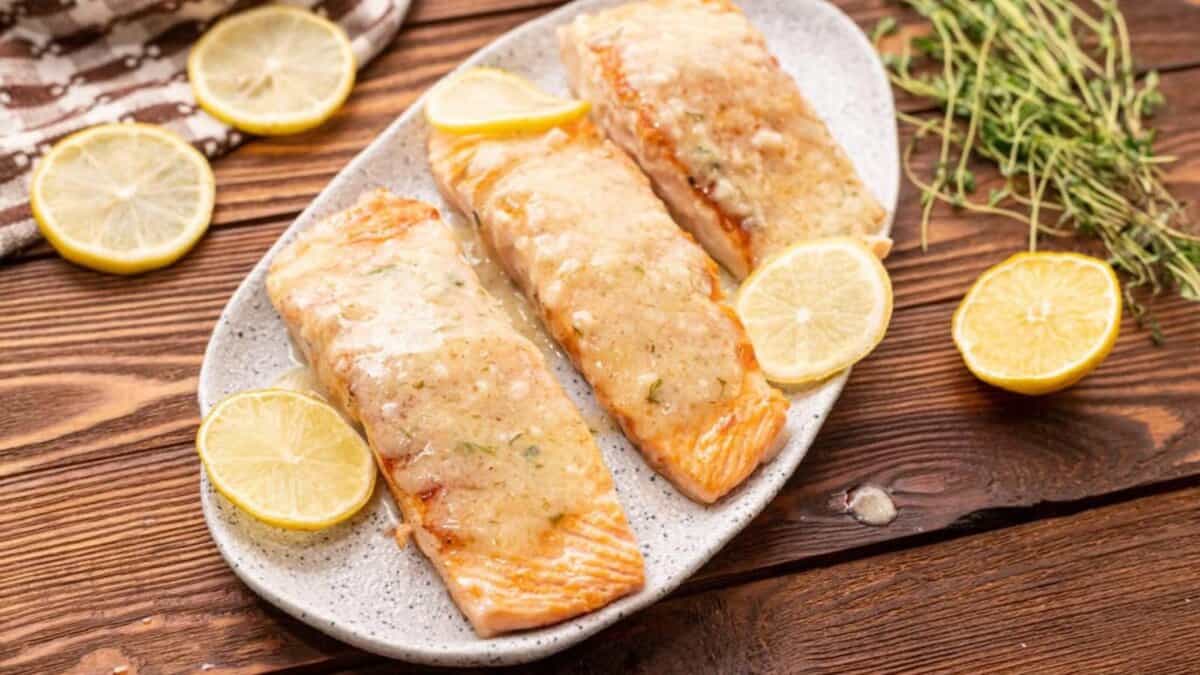 Three cooked salmon fillets topped with a creamy sauce are arranged on a plate, garnished with lemon slices. Fresh herbs and more lemon slices are on the rustic wooden table beside the plate.